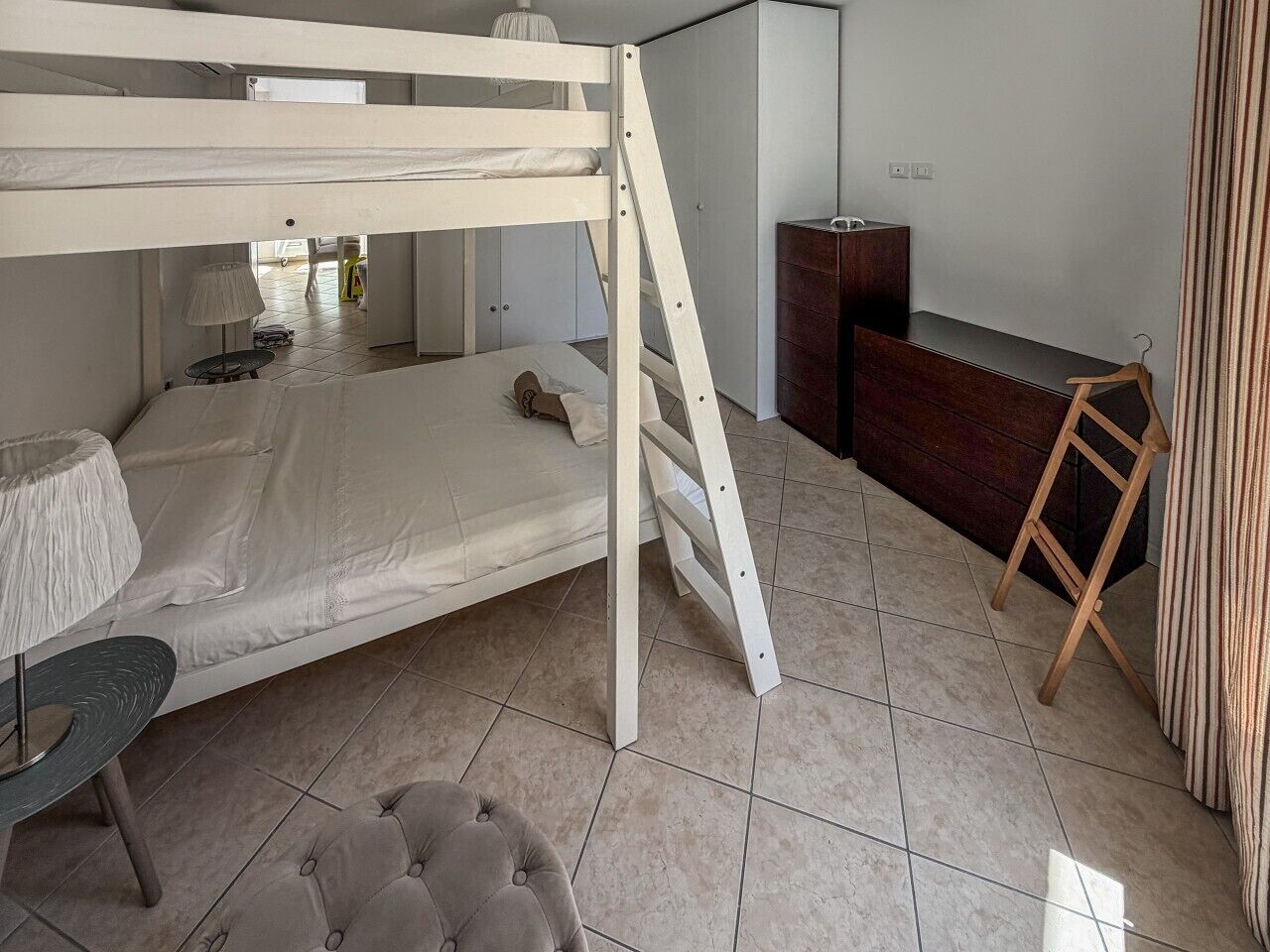 A bunk bed with a ladder, a desk with a lamp, a chair, a dresser, and a striped curtain.