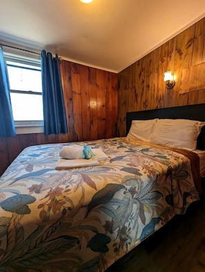 Ein Zimmer mit Holzwandverkleidung, einem Bett mit Blumenmuster auf der Bettwäsche, einem Fenster mit Vorhängen und einer wandmontierten Leuchte.