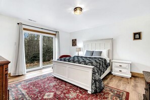 Ein Schlafzimmer mit Bett, Nachttisch, Lampe, einem Fenster mit Vorhängen und Blick auf Bäume.