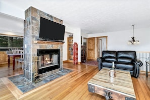 A living room with a stone fireplace, a black leather sofa, and a flat-screen TV mounted above the fireplace. - Charming Mountain Treehouse with Forest Views near Downtown Black Mountain, NC (Black Mountain)