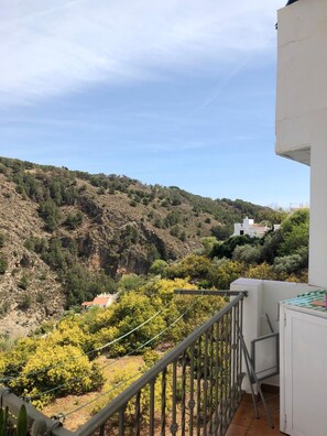 Ein Balkon mit Blick auf eine hügelige Landschaft, ein weißes Gebäude und ein klarer Himmel.