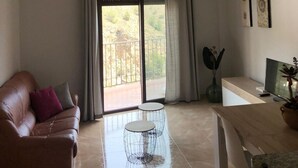 Una sala de estar con un sofá de cuero, dos mesitas de noche de metal, una mesa de centro de madera, un televisor y un ventanal corredizo que da a un balcón.