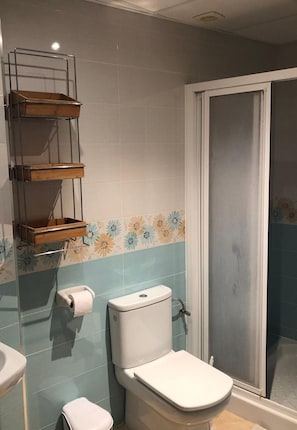 Ein Badezimmer mit Toilette, Toilettenpapier, einer Dusche mit Glastür und einem Regal mit einer Holztablett.