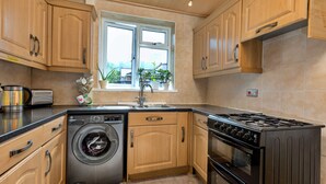 A kitchen with wooden cabinets, a washing machine, a stove, and a window with plants. - Oakdene (Stockton-on-Tees)