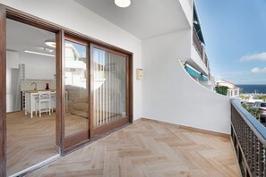 Uma varanda com vista para o mar, uma porta de correr e uma cozinha, e um piso com padrão em espinha de peixe.