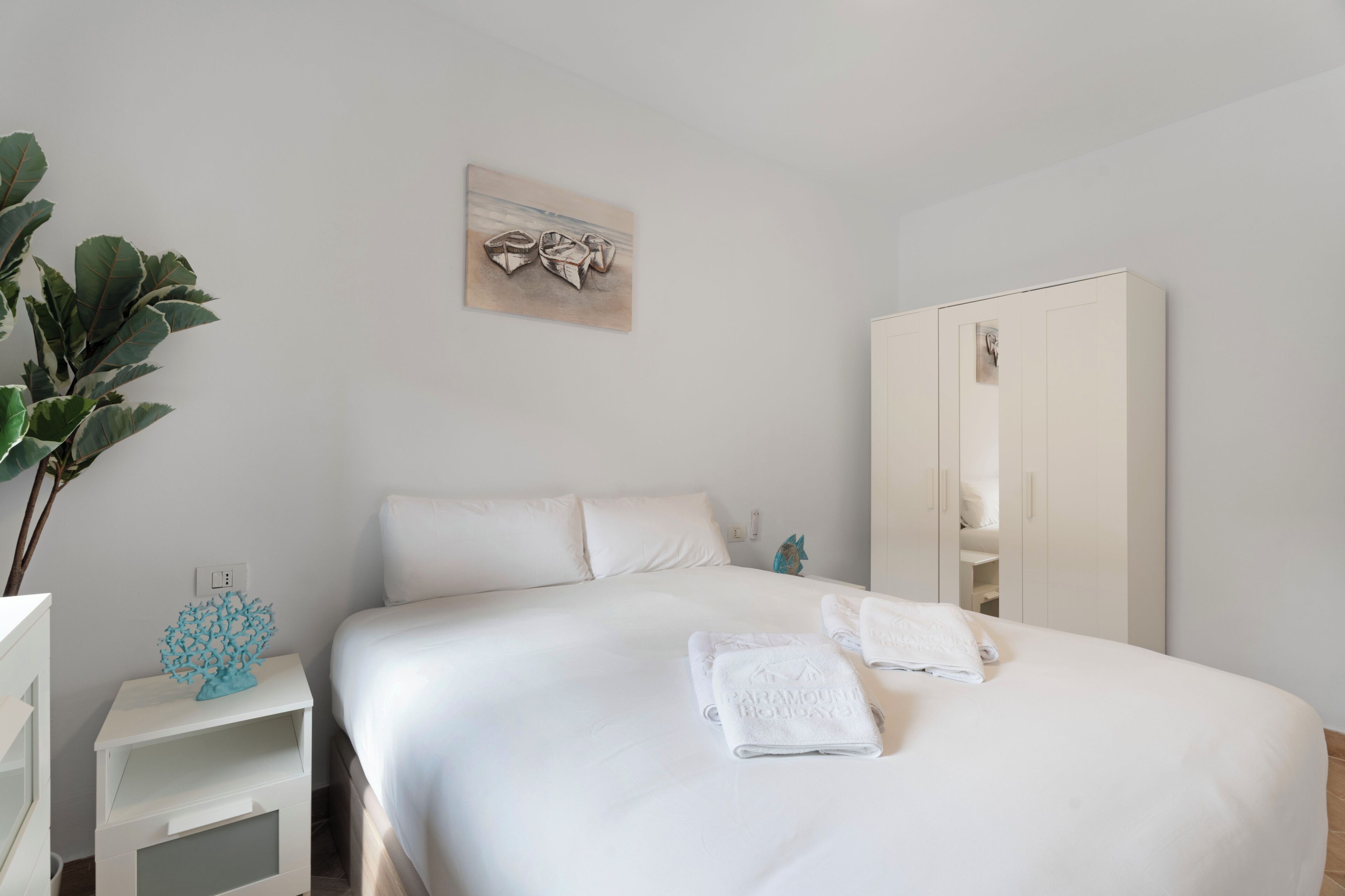 A neatly made bed with white linens, a white wardrobe, and a wall-mounted artwork.