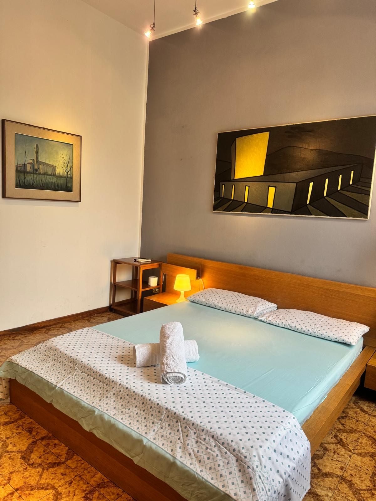 A hotel room with a bed, a bedside table, a lamp, a painting, and a view of a building.