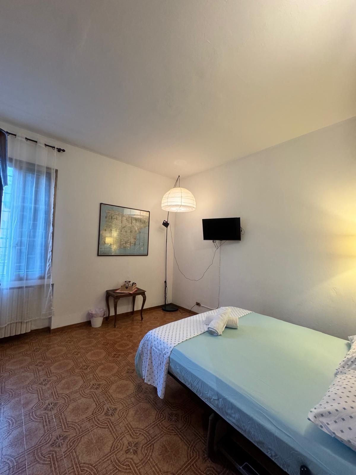 Una camera con un letto, una scrivania, una sedia, una lampada da terra e una TV a parete.