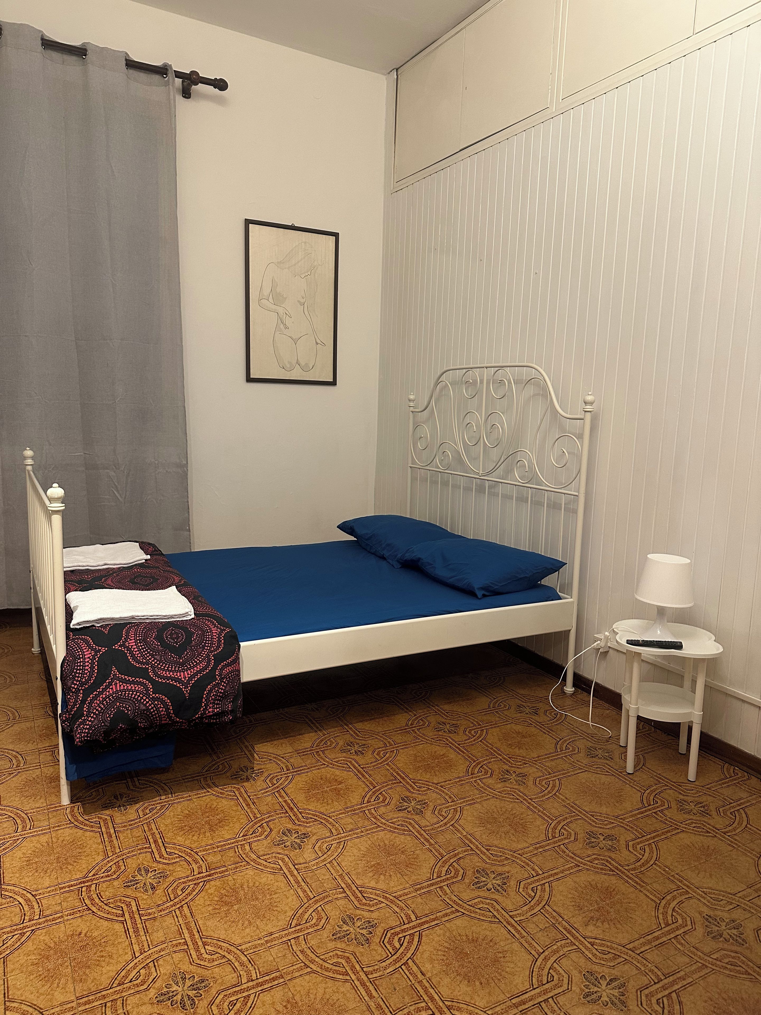 Una cama individual con colchón azul, cabecera de metal blanca, mesita de noche con lámpara y suelo estampado.