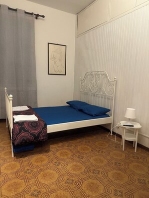 A single bed with a blue mattress, a white metal headboard, a bedside table with a lamp, and a patterned floor.