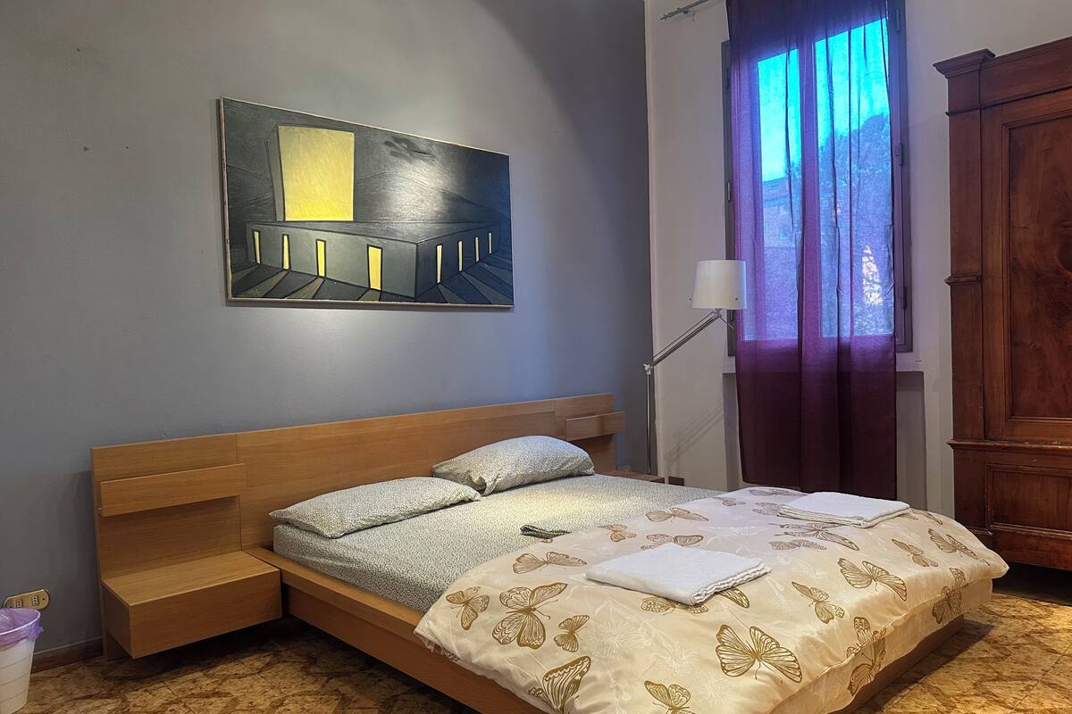 A bedroom with a bed, a nightstand, a lamp, a window with curtains, a wooden cabinet, and a painting on the wall.