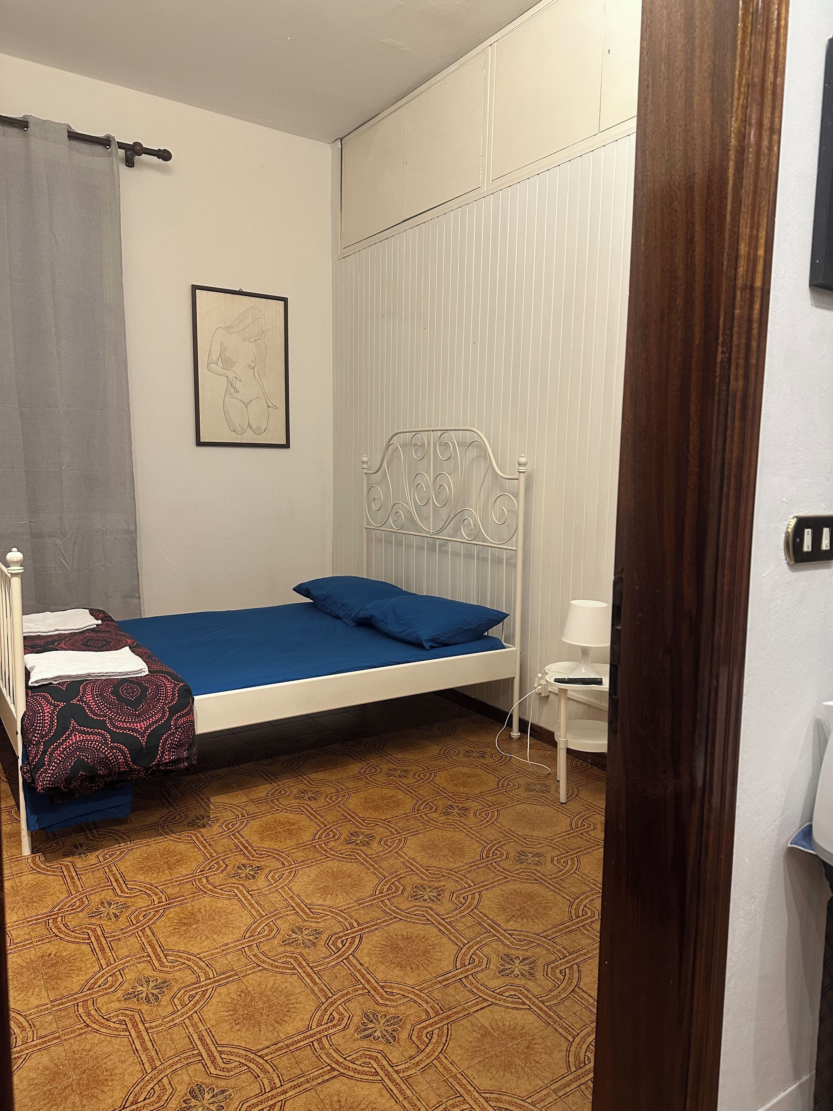 Ένα μονό κρεβάτι με ένα μπλε στρώμα, ένα λευκό μεταλλικό προσκέφαλο, μια γκρι κουρτίνα, μια κορνιζαρισμένη εικόνα στον τοίχο και ένα δάπεδο με σχέδια.