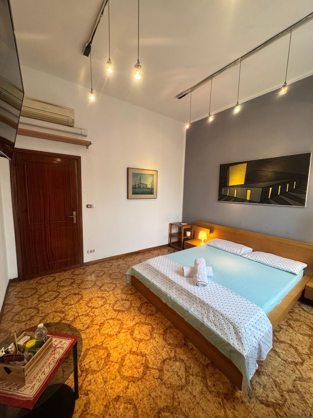 A hotel room with a bed, a small table with a tray, a painting on the wall, and a ceiling light fixture.