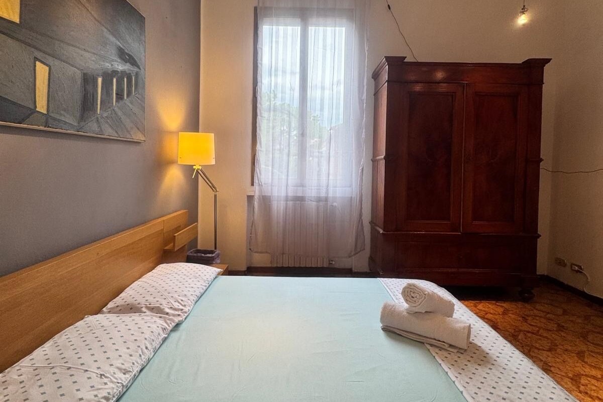 A bedroom with a bed, a wooden wardrobe, a painting on the wall, and a window with curtains.