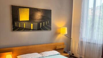 A hotel room with a bed, a nightstand, a lamp, a painting on the wall, and a window with curtains.