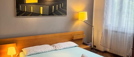 A hotel room with a bed, a nightstand, a lamp, a painting on the wall, and a window with curtains.