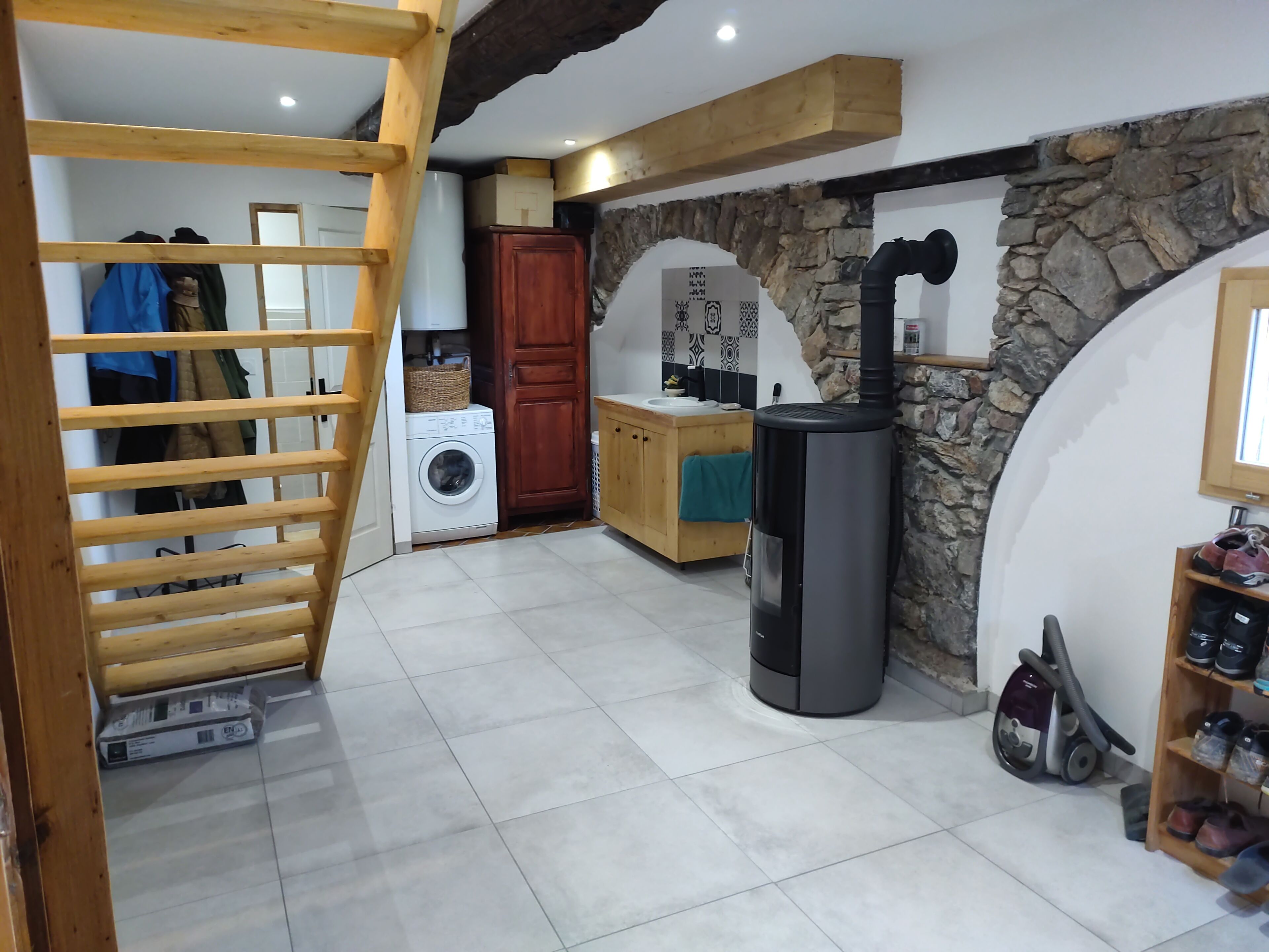 Een wasruimte met een wasmachine, een droogrek, een houten trap, een stenen muur en een houtkachel.