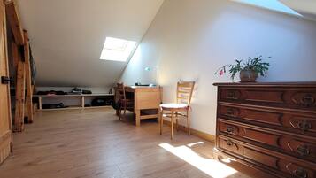 Une chambre mansardée avec un plafond en pente, un plancher en bois, un bureau avec une chaise, une commode et une plante en pot.