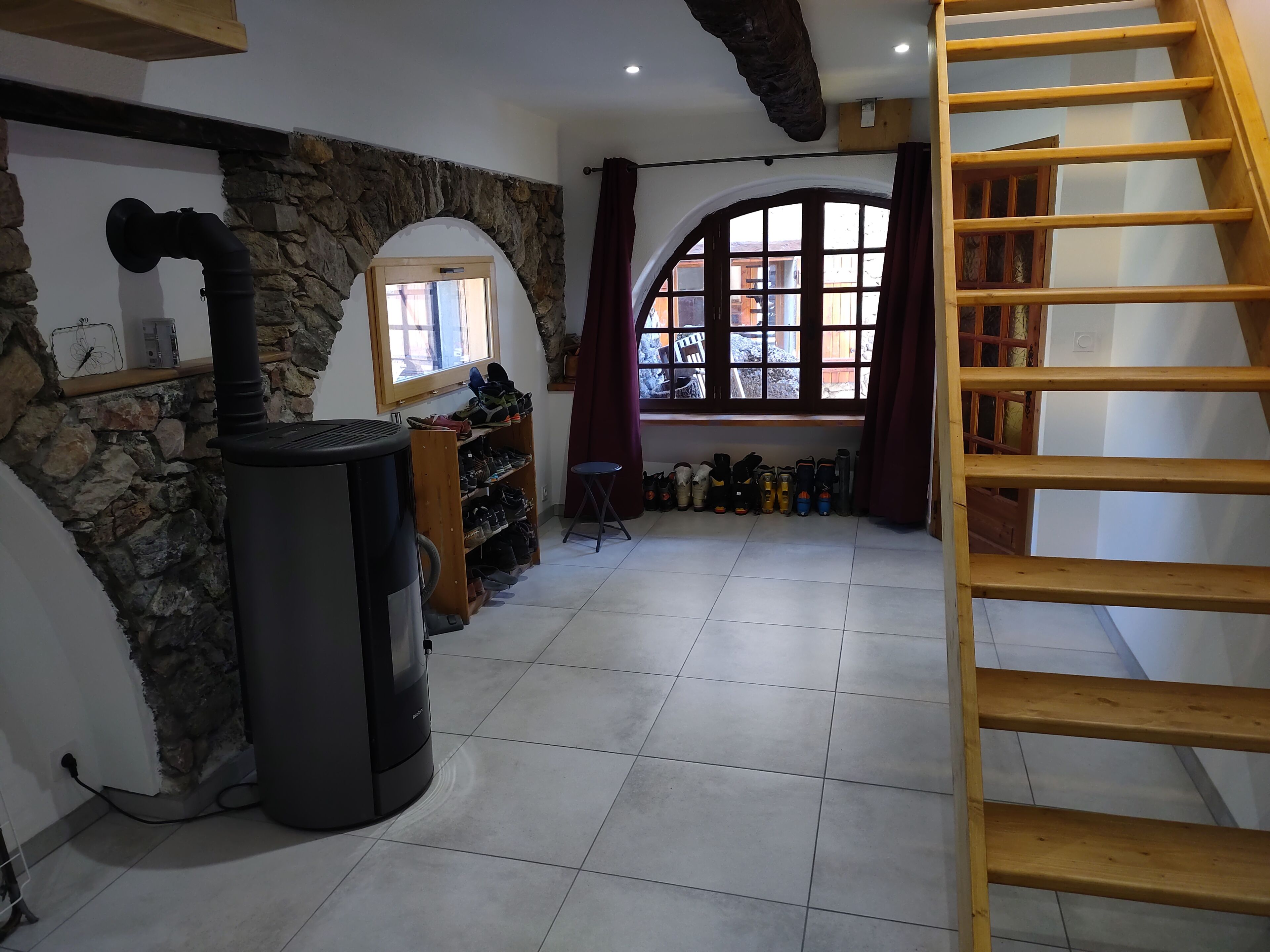 Une pièce avec un mur en pierre, un radiateur noir, une étagère en bois où sont rangées des chaussures, une fenêtre avec des rideaux et un escalier en bois.