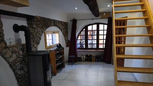 A room with a stone wall, a black heater, a wooden shelf with shoes, a window with curtains, and a wooden staircase.