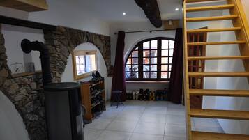 Une pièce avec un mur en pierre, un radiateur noir, une étagère en bois où sont rangées des chaussures, une fenêtre avec des rideaux et un escalier en bois.