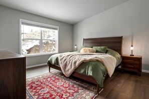 Ein Schlafzimmer mit Bett, Nachttisch, Lampe, Fenster und einem gemusterten Teppich.