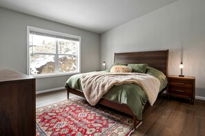 Quarto com cama, mesa de cabeceira, abajur, janela e tapete estampado.