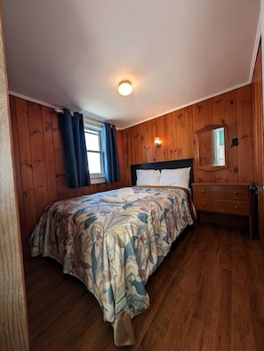 Ein Schlafzimmer mit Holzwänden, einem Bett mit floralem Bettbezug, einem Nachttisch, einer Kommode und einer Deckenleuchte.