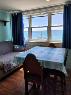 Ein Zimmer mit Bett, Tisch mit Stühlen, einem Fenster mit Vorhängen und Blick aufs Meer.