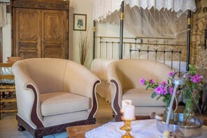 Ein Zimmer mit zwei Sesseln, einem Bett mit Metallkopfteil, einem Holzschrank und einer Vase mit Blumen.