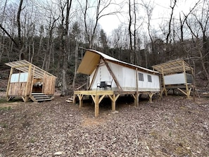 Three wooden cabins with elevated platforms, a table, and chairs in a forested area.