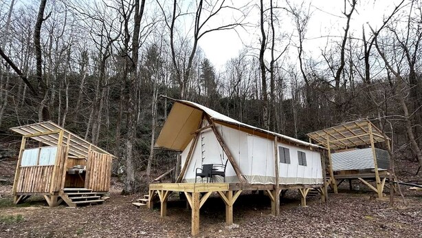 Three wooden cabins with elevated platforms, a table, and chairs in a forested area.