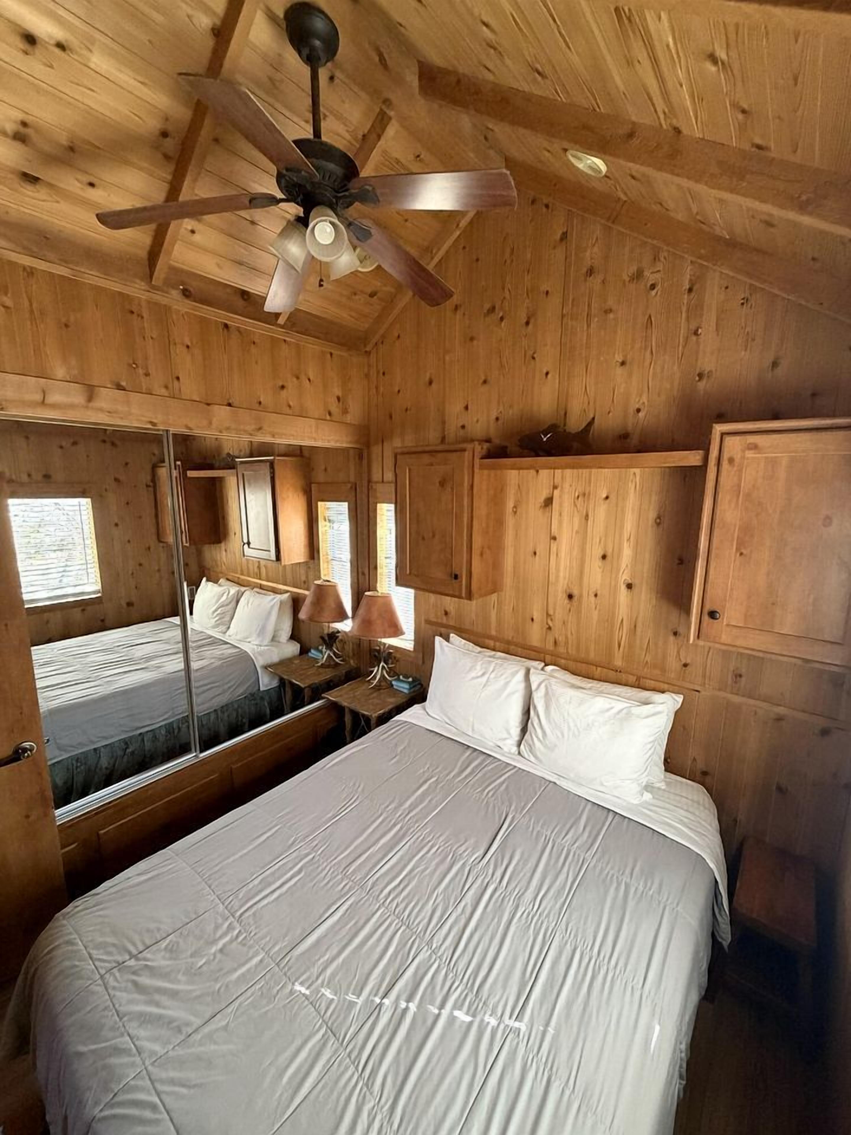 A wooden cabin room with a bed, a ceiling fan, a mirror, and a window.