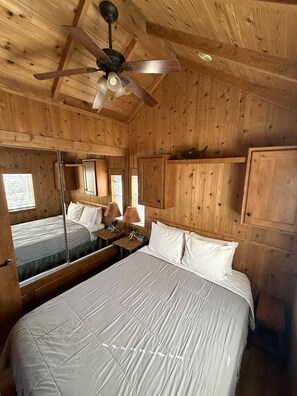Ein Zimmer einer Holzhütte mit einem Bett, einem Deckenventilator, einem Spiegel und einem Fenster.