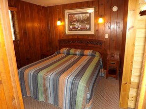 Ein Zimmer mit Holzwand, einem Bett, einem gerahmten Bild an der Wand und einem kleinen Nachttisch.