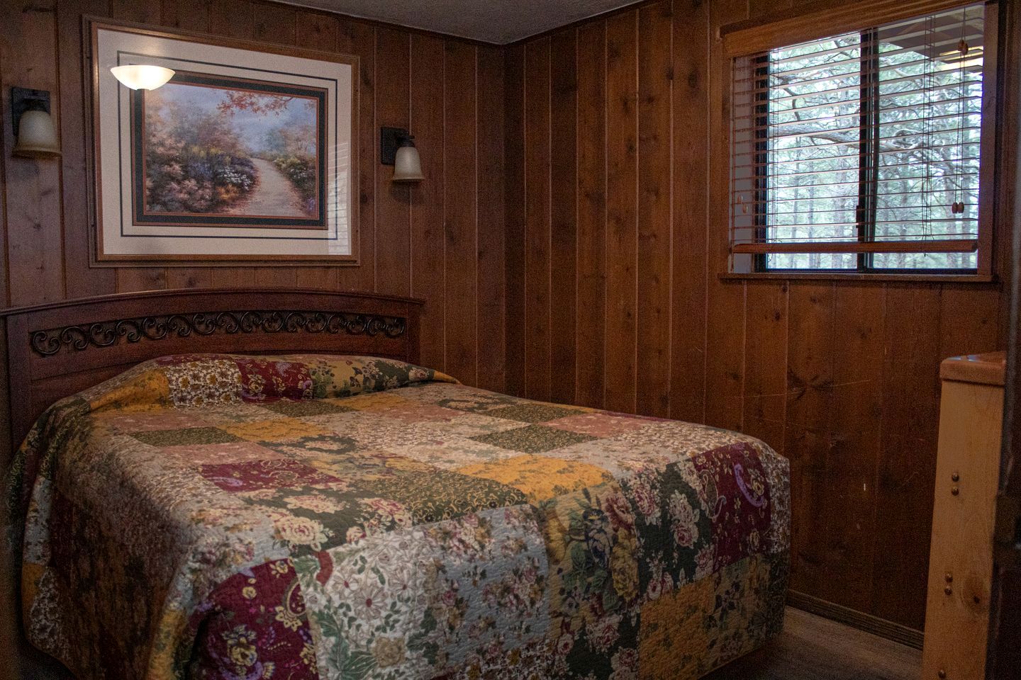 A bedroom with a wooden paneled wall, a bed with a floral quilt, a framed painting above the bed, and a window with blinds.