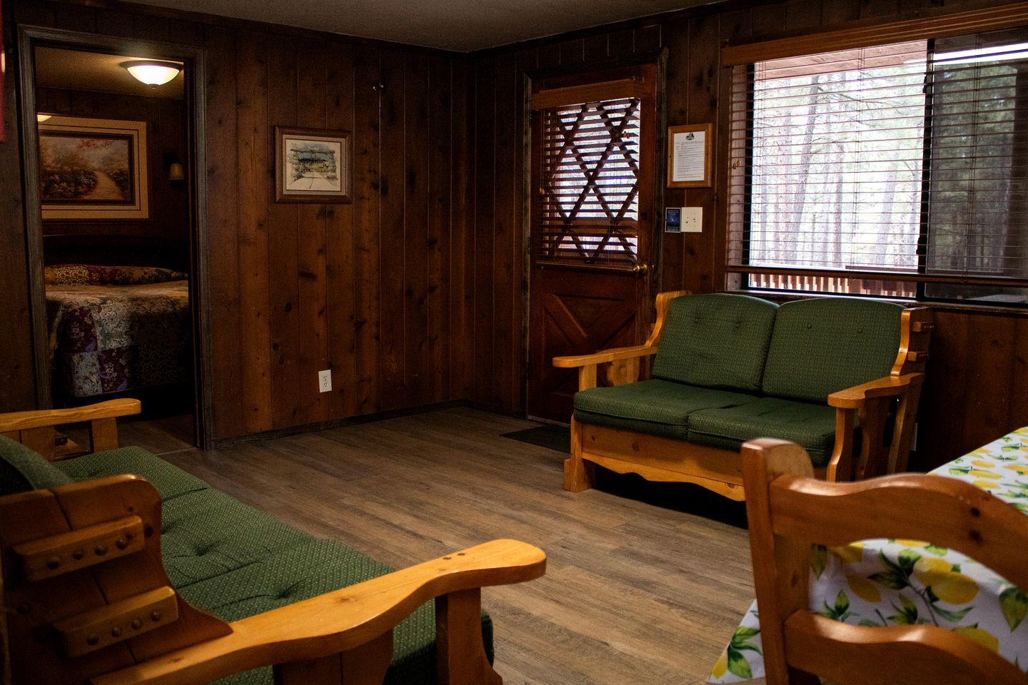 A wooden-floored room with a green sofa, wooden chairs, and a bed with a patterned blanket.