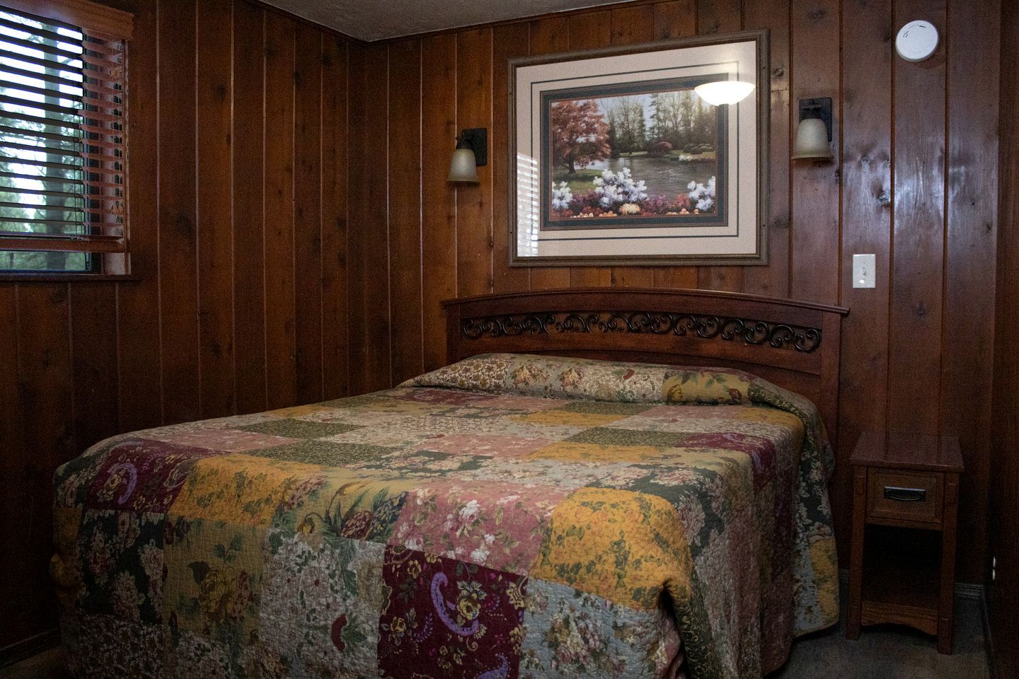 A bedroom with a wooden paneled wall, a bed with a floral quilt, a framed picture above the bed, and a nightstand with a lamp.