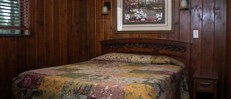Ein Schlafzimmer mit einer holzverkleideten Wand, einem Bett mit floralem Patchwork, einem gerahmten Bild über dem Bett und einem Nachttisch mit Lampe.