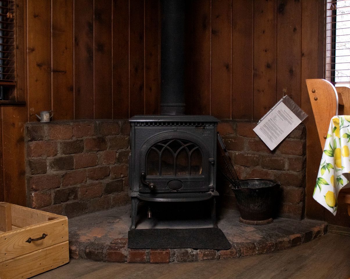 A wood-burning stove with a chimney, a brick fireplace, a wooden chest, and a metal bucket.