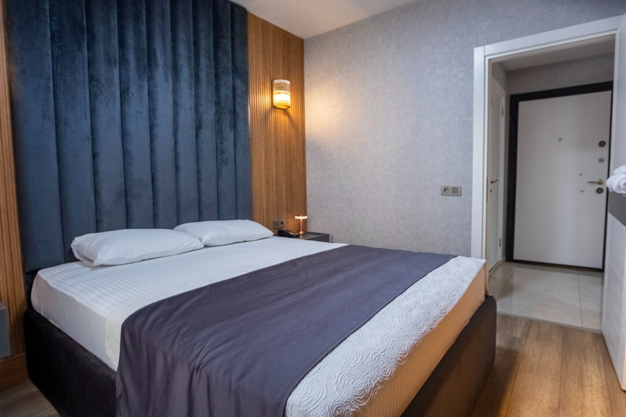 Una habitación de hotel con cama, mesita de noche, lámpara de pared y una puerta.