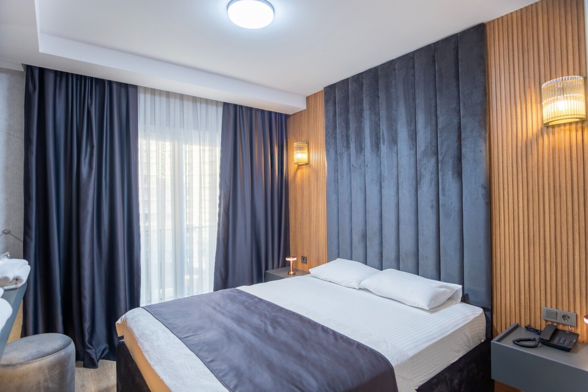 Habitación de hotel moderna con una cama grande, una mesita de noche con lámpara, un escritorio pequeño con teléfono y una ventana con cortinas.
