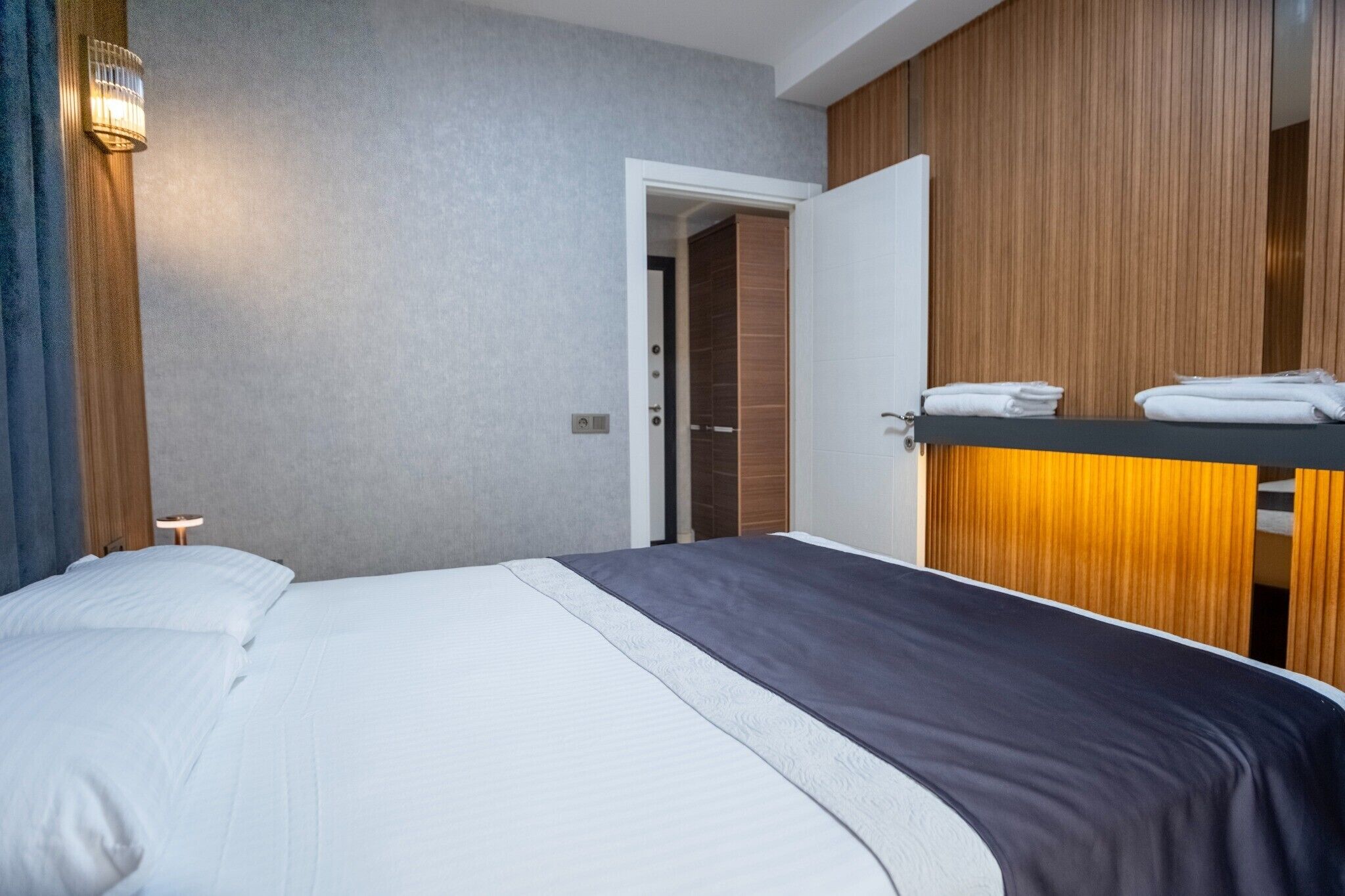 Une chambre d’hôtel comprenant un lit, une table de chevet, une étagère fixée au mur avec des serviettes, et une porte donnant sur une autre pièce.