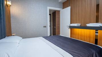 Une chambre d’hôtel comprenant un lit, une table de chevet, une étagère fixée au mur avec des serviettes, et une porte donnant sur une autre pièce.