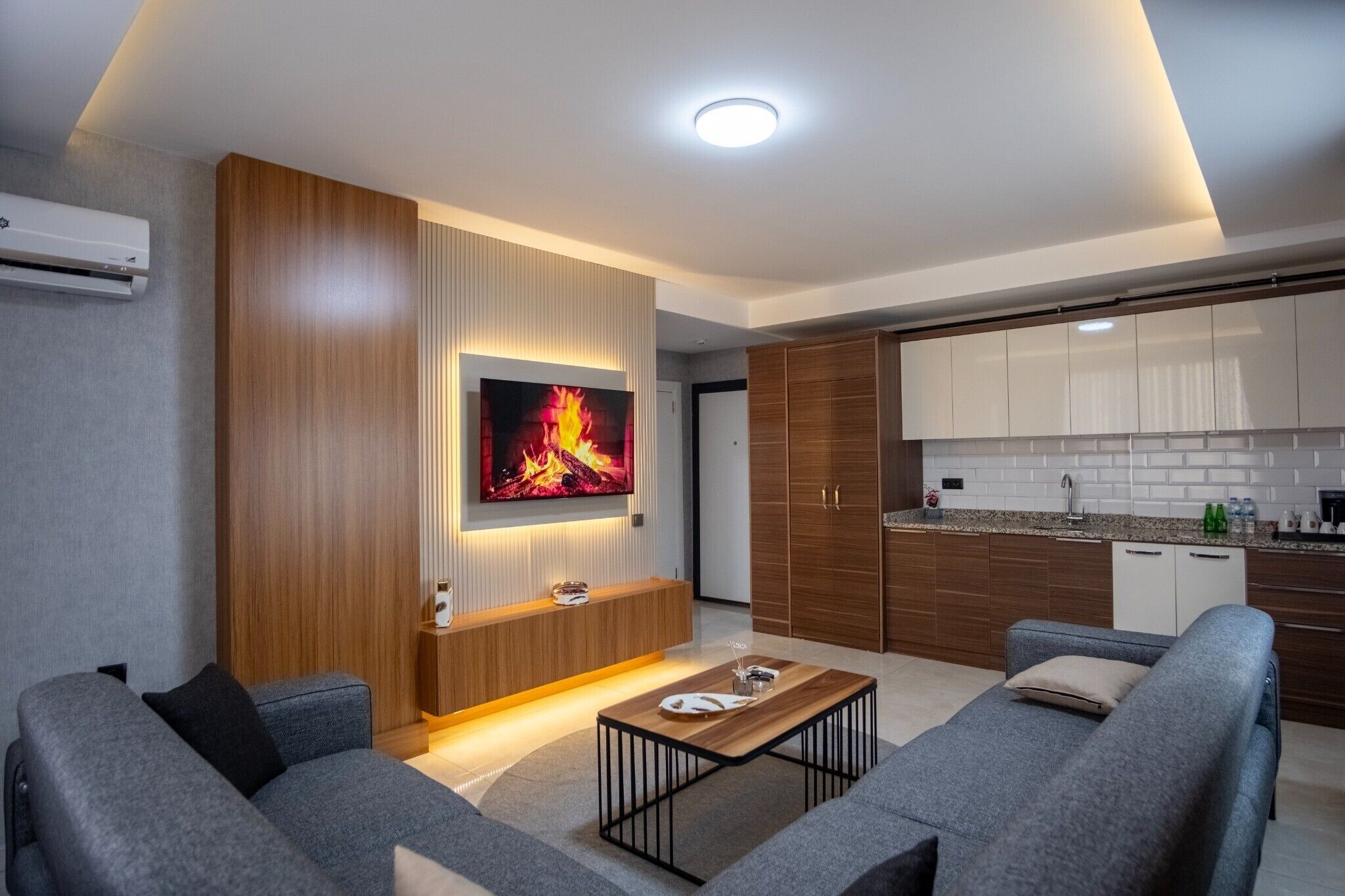 A modern living room with a flat-screen TV, a wooden entertainment unit, grey sofas, and a kitchen area with wooden cabinets.
