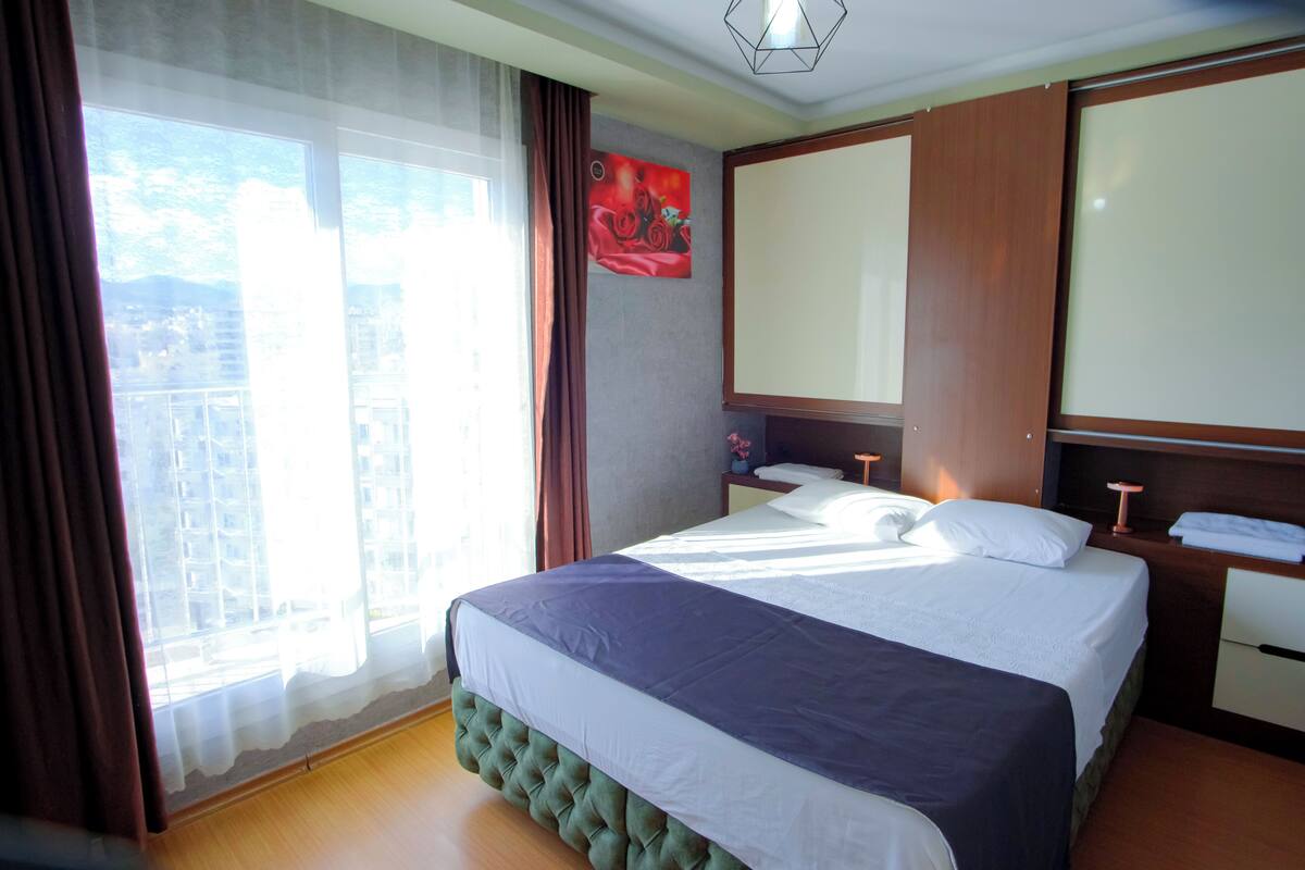 A hotel room with a large bed, a bedside table, a mirror, and a window with curtains.