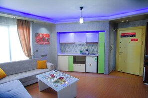 A compact hotel room with a kitchenette, a sofa with cushions, a coffee table with a magazine, and a door with a sign.