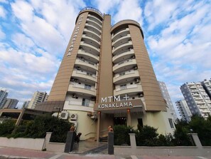Um hotel de vários andares com varandas, uma entrada central e céu claro.