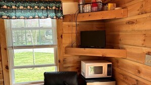 A compact room with wooden walls, a window with a view, a microwave, a refrigerator, a chair, and a trash bin.