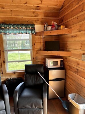 A compact room with wooden walls, a window with a view, a microwave, a refrigerator, a chair, and a trash bin.